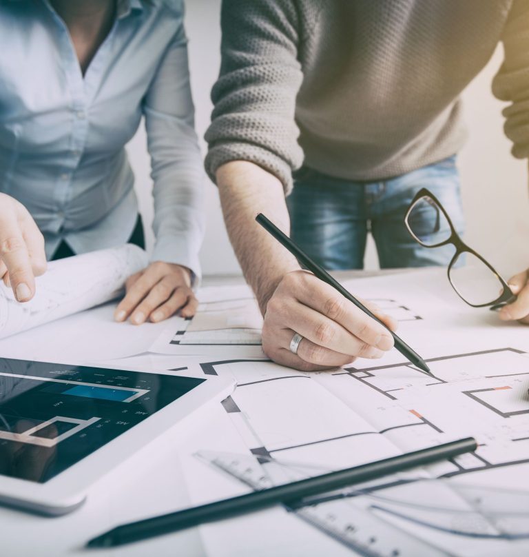 Duo di architetti lavora su planimetrie con dispositivi digitali e strumenti di progettazione su un tavolo