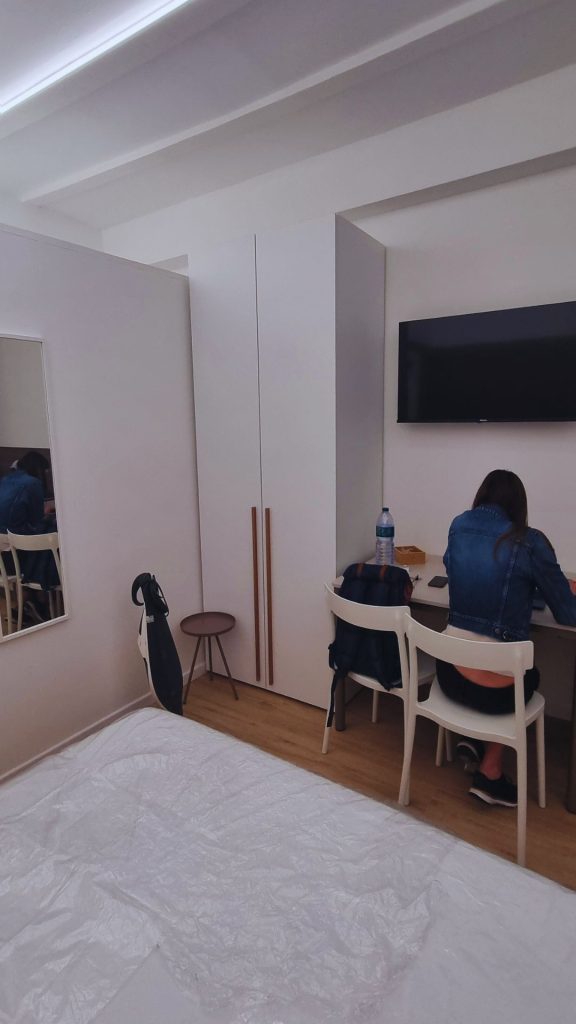 Vista dal letto sul lato opposto del monolocale: armadio a tutta altezza, postazione smart working integrata con TV a parete e sedia da pranzo, ideale per affitto breve.