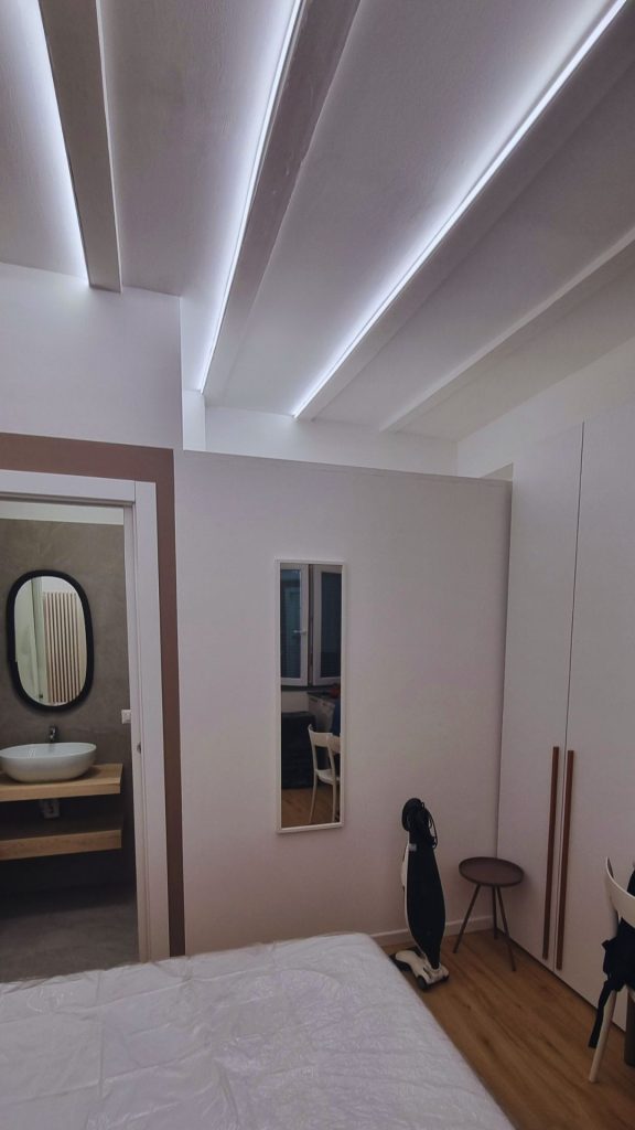 Vista dalla zona letto verso il bagno, con illuminazione a LED integrata nel soffitto e arredi su misura per ottimizzare lo spazio. A sinistra si nota l’accesso al bagno con lavabo d'appoggio e specchio ovale.