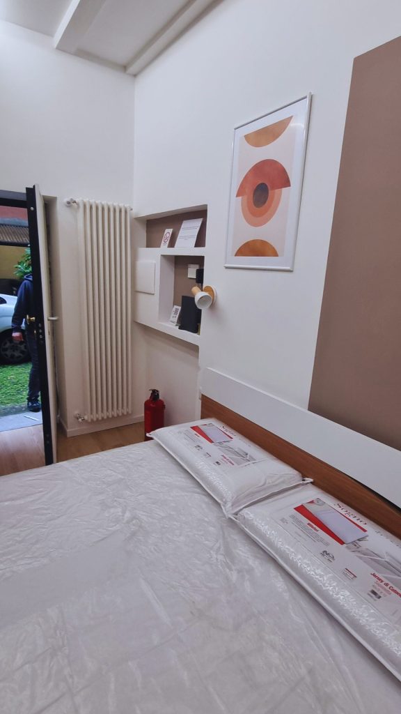 Vista interna del monolocale di 20 mq con letto matrimoniale, testiera integrata e parete attrezzata con nicchie funzionali, quadro decorativo e accesso diretto al giardino condominiale.