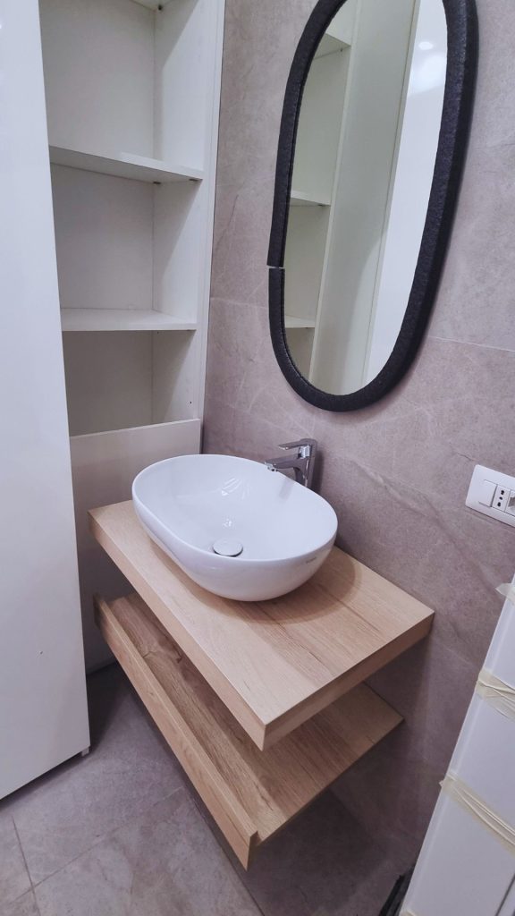 Mobile bagno sospeso con lavabo da appoggio ovale, mensola in legno e specchio a cornice nera, in ambiente compatto ed essenziale.