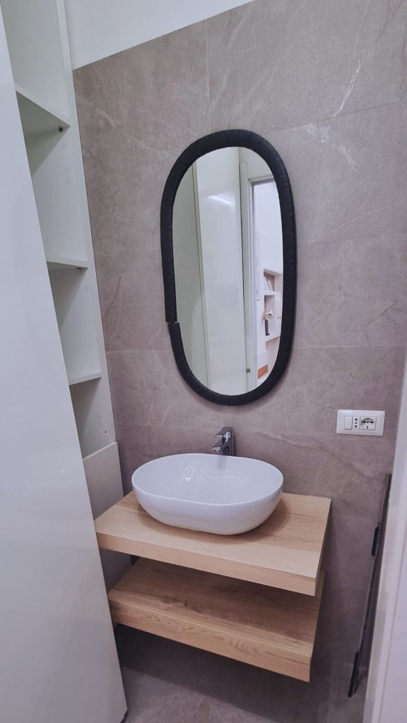 Vista frontale del bagno compatto con lavabo da appoggio su mensola in legno, specchio ovale a cornice nera e parete rivestita in gres effetto pietra.