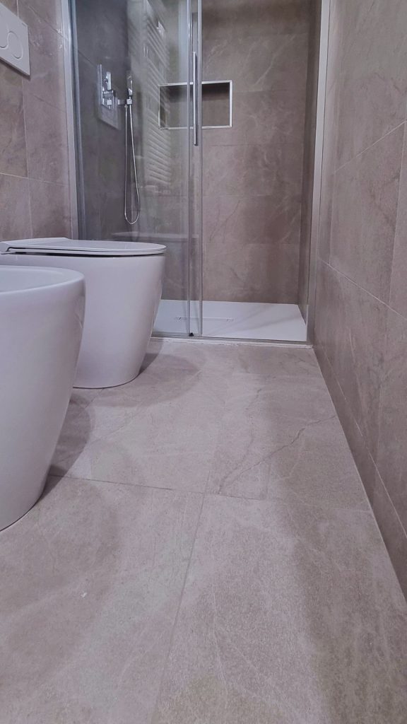 Bagno compatto del monolocale ristrutturato a Milano, con sanitari filomuro sospesi, doccia con piatto filo pavimento e nicchia integrata nel rivestimento in gres effetto pietra.