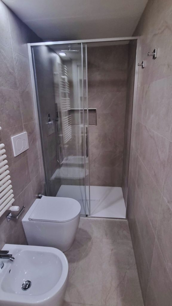 Vista completa del bagno ristrutturato in monolocale da 20 mq, con sanitari sospesi, doccia in nicchia con vetro scorrevole e rivestimento uniforme in gres effetto pietra per ottimizzare lo spazio e garantire continuità visiva.